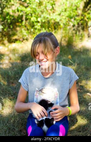 Defocus blonde little smiling girl playing with cat, black and white small kitten. Nature green summer background. Girl stroking pet. Love animals Stock Photo