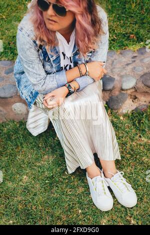 Seated modern independent woman, with pink hair, dark glasses, silver-gold skirt and white sneakers. Hipster woman Stock Photo