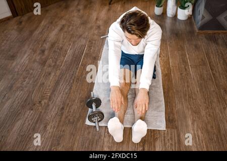 Focused sportive male sitting on the floor, doing stretching warming up before workout. Handsome man training at home while quarantine, keeping Stock Photo