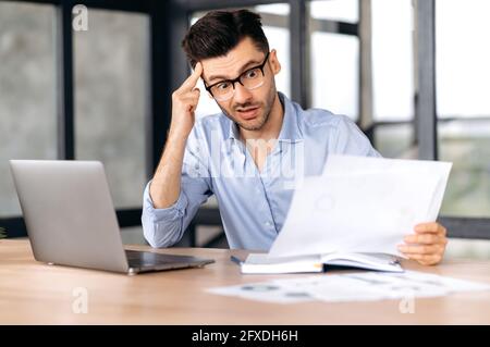 Shocked frustrated young caucasian business leader, top manager or freelancer wearing glasses sits at the workplace in the office, looks with surprise at business documents, puzzled Stock Photo