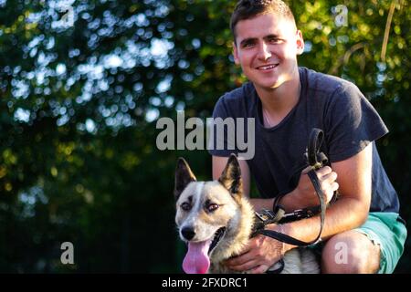 Defocus close-up young caucasian man smiling and seating with dog. The portrait of a guy squatting near the siberian laika husky outdoor at summer Stock Photo