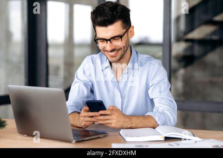 Joyful caucasian young man, manager or freelancer sitting at his desk in the office, texting with a friend or client using a smartphone, online chat, browsing the Internet, answering email, smiling Stock Photo