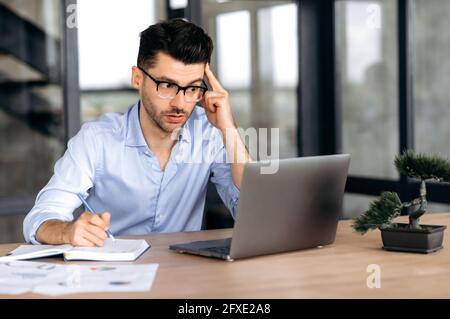 Focused pensive intelligent caucasian manager, freelancer or ceo, wearing glasses, sitting at table in modern office, using laptop, going through online training, brainstorming, takes notes Stock Photo