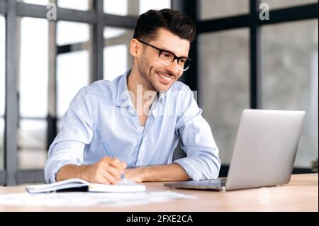 Handsome successful caucasian manager or freelancer wearing glasses, sitting at workplace in office, using laptop, taking notes, studying or working remotely, listening to webinar, smiling Stock Photo