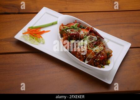 Chilli Chicken dry,well garnished and arranged in an oval shaped bowl on a white serving plate with wooden texture Stock Photo