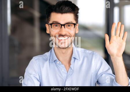 Caucasian handsome confident successful businessman, coach or consultant in glasses, communicates via video conference with client or employees, greets with hand gesture, looks at webcam, smiles Stock Photo