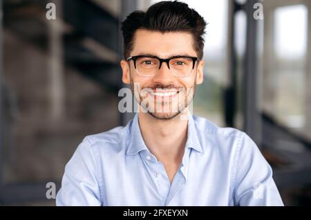 Close up portrait of handsome confident successful young caucasian man wearing glasses, wearing stylish shirt, sitting at office, looking directly at camera and smiling friendly Stock Photo