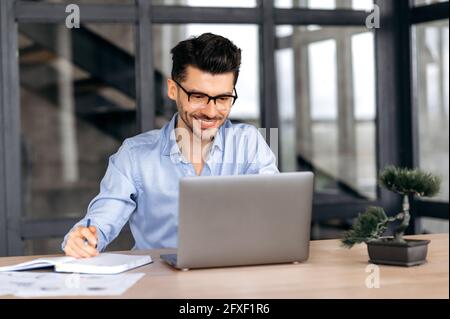 Confident joyful attractive young caucasian businessman, broker or manager in formal clothes and glasses sitting at table in office, taking notes, using laptop, looking at screen, smiling Stock Photo