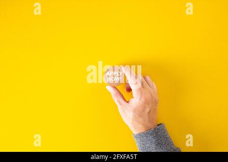 Male hand holds a wooden block with the word apply now on yellow background. Job vacancy, education or membership application concept. Stock Photo