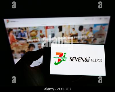 Person holding cellphone with logo of Japanese retail company Seven and i Holdings Co. Ltd. on screen in front of website. Focus on phone display. Stock Photo