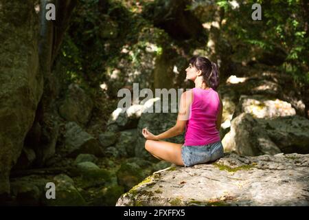 A woman sits in a lotus field on a large rock among the rocks in the open air and meditates, enjoys the unity with nature, listens to the silence and Stock Photo