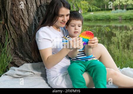 Mom and baby boy 4 years old play with a colored trendy toy Pop it in the park in nature. Antistress sensitive toy or reusable bubble wrap. Trend of 2 Stock Photo