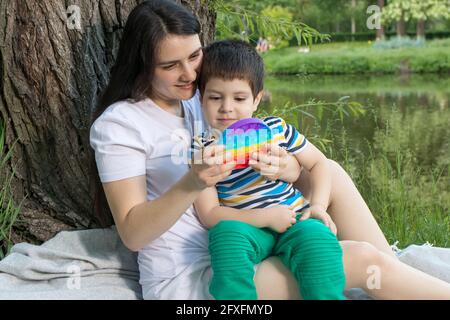 Mom and baby boy 4 years old play with a colored trendy toy Pop it in the park in nature. Antistress sensitive toy or reusable bubble wrap. Trend of 2 Stock Photo