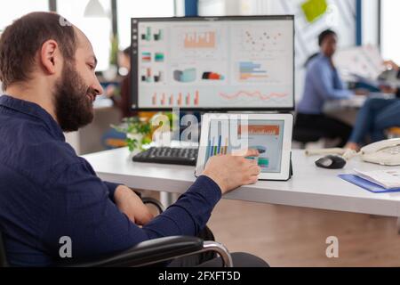 Paralysed business man sitting immobilized in wheelchair working at tablet and computer in same time analysing financial graphs, pc showing data prosessing on display for project regarding economy. Stock Photo