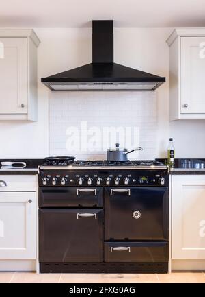 Black enamel oven range cooker with chimney hood in a modern kitchen with shaker style painted wood units in neutral off white. UK farmhouse kitchens Stock Photo