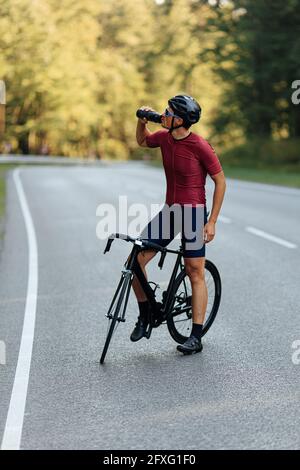 Strong man drinking water while standing on road with bike Stock Photo