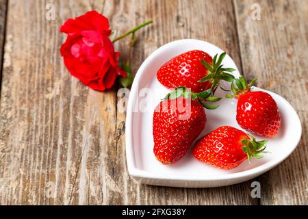 Ripe strawberries in a heart shaped plate heart-shaped plate and rose on a wooden background. Still life with delicious berries. Stock Photo