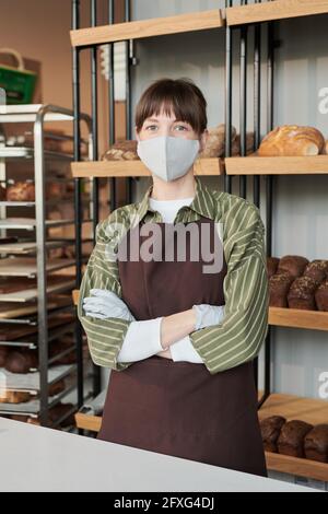 Portrait of young saleswoman in uniform and mask standing in bakery shop Stock Photo