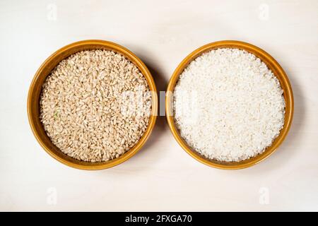 top view of polished and unpolished medium-grain rice in round ceramic bowls on light brown wooden table Stock Photo