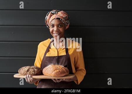 Portrait of young baker in apron holding the tray with fresh bread and smiling at camera Stock Photo