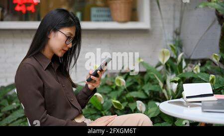 Side view of female office worker using smartphone while siting and waiting for coffee in coffee shop Stock Photo