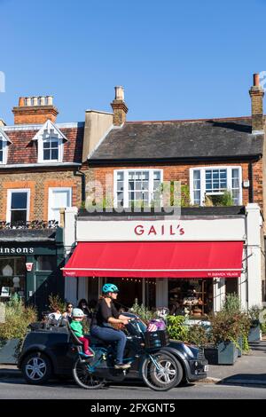 A young woman transports her child by bicycle past Gail's bakery in Dulwich Village, London, England, UK. Stock Photo