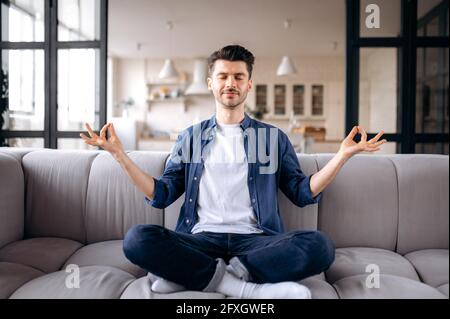 Relaxed attractive young caucasian man, calm pacified guy in casual clothes sits on the sofa in the living room with closed eyes, meditates in the lotus position, relieves stress, resting Stock Photo