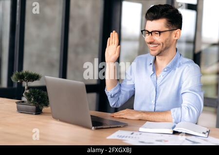 Cute friendly young Caucasian businessman, manager or freelancer wearing glasses, sitting at a laptop, communicating via video conference with friends or colleagues, gesturing with his hand, smiling Stock Photo