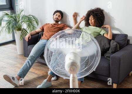 exhausted interracial couple sitting on couch near blurred electric fan in living room Stock Photo