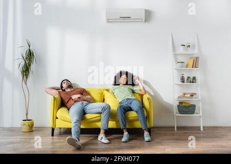 exhausted multiethnic couple sitting on yellow sofa and suffering from heat in summer Stock Photo