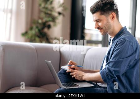 Concentrated young adult Caucasian man, freelancer or student, in casual clothes, sitting on sofa in living room, using laptop for video calling with colleagues, looking at screen, taking notes Stock Photo