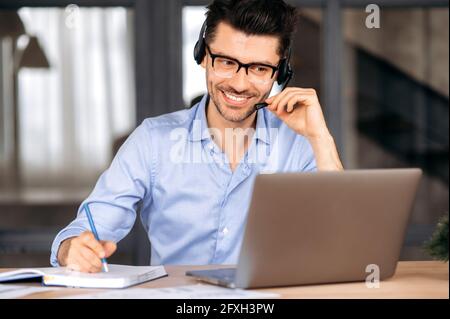 Joyful confident young caucasian man wearing glasses and headset, freelancer, office worker or consultant, using laptop for online consultation or negotiation, communicating via video call, smiling Stock Photo