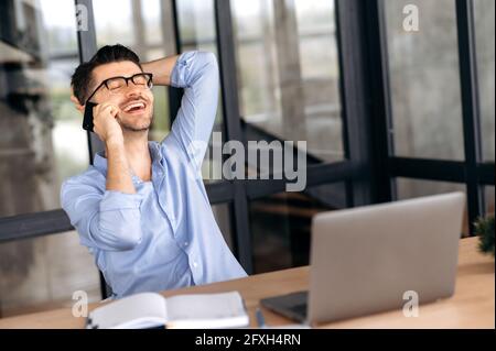 Happy friendly successful caucasian guy with glasses, office worker, manager or broker, sitting at the table in the office, leaning back, talking on the phone with friends, taking a break, smiling Stock Photo