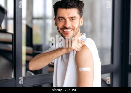 Vaccination and treatment during a coronavirus or flu pandemic. Satisfied young caucasian man, wearing casual clothes, with adhesive plaster on shoulder after vaccination, looking at camera, smiling Stock Photo