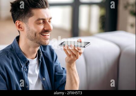 Peaceful relaxed young attractive caucasian man in stylish clothes sitting on the couch and recording voice message or talking on smartphone on speakerphone with friends or family, closing his eyes Stock Photo
