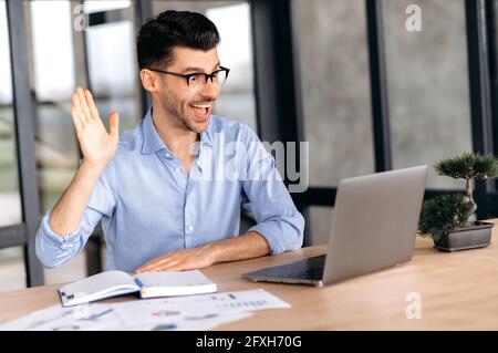 Attractive successful cute young caucasian broker, top manager or freelancer wearing glasses sitting at the table chatting online with a client or colleagues, greeting with a hand gesture, smiling Stock Photo