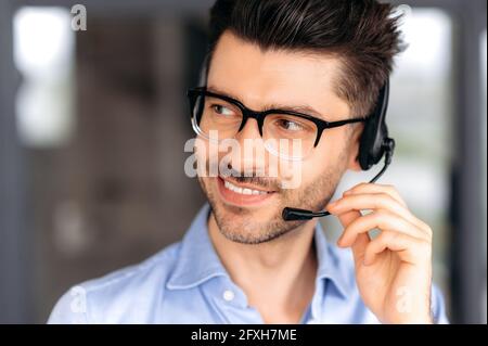 Close up portrait of successful joyful young caucasian operator or call center manager wearing glasses, in formal attire, using headset for video conference or phone call, looking away, smiling Stock Photo