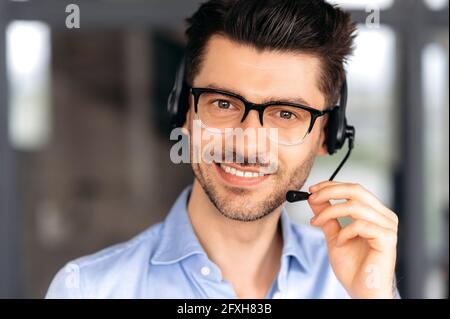 Close up portrait of attractive happy young caucasian businessman, call center operator or manager wearing glasses, uses headset for conference calling, looking at camera and smiling friendly Stock Photo