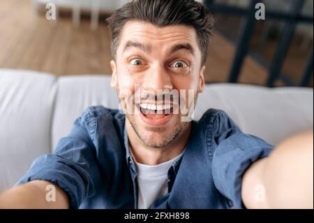 Portrait of happy young caucasian man in casual clothes sitting on sofa taking selfie, grimacing while shooting, holding smartphone in hands and looking at camera enthusiastically, smiling Stock Photo