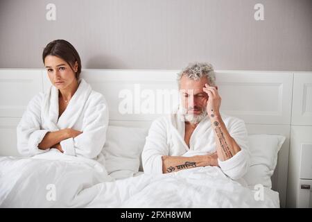 Exited brunette woman and thoughtful bearded man in white bathrobes looking away in bedroom Stock Photo