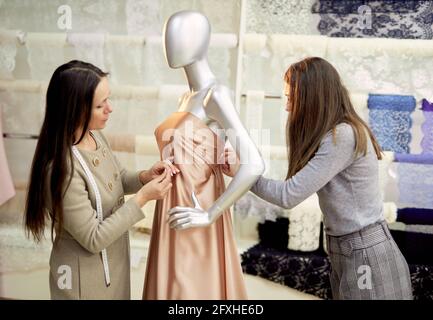 Professional brunette team of tailors, dressmakers or designers working with new model applying linen fabrics cloth on mannequin or dummy in atelier. Fashion and tailoring concept. High quality image Stock Photo