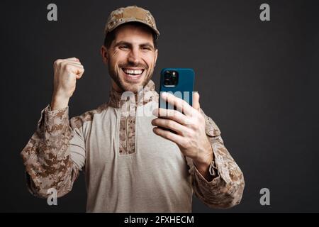 Excited military man making winner gesture while using cellphone isolated over black background Stock Photo