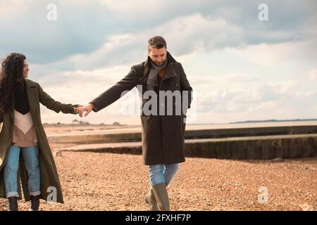 Couple in winter coats holding hands walking on sunny beach Stock Photo