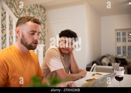 Gay male couple eating and using laptop in kitchen Stock Photo