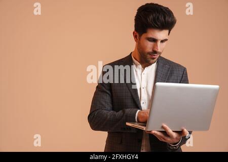 Serious young man in jacket standing while working with laptop isolated over beige background Stock Photo
