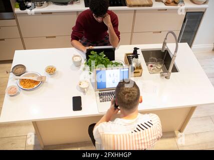 Young gay male couple working from home at laptops in kitchen Stock Photo