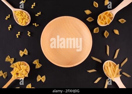 Different pasta types in wooden spoons and empty wooden plate on black table. Top view. Stock Photo
