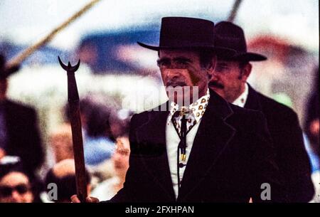 A gardian wearing a black hat and carrying a trident on horseback amidst the throng at the Romani pilgrimage in Saintes-Maries-de-la-Mer; in the background, a second man in similar traditional dress, May 1978. Saintes-Maries-de-la-Mer, Arles, Bouches-du-Rhône, Provence-Alpes-Côte d'Azur, 13460, France Stock Photo