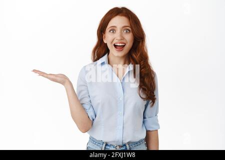 Excited and amazed redhead woman holds product in open hand, holding something cool, showing at item in palm, standing in blouse against white Stock Photo