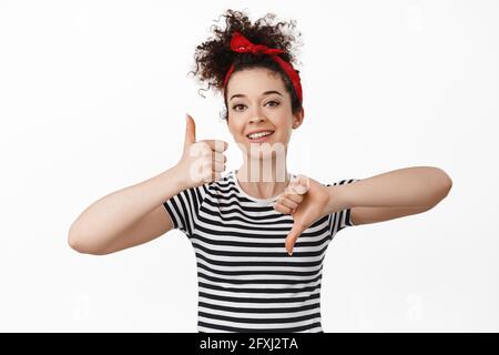 Medium. Smiling brunette woman showing thumbs up and down, rating and feedback concept. standing in headband with combed curly hair and t-shirt Stock Photo